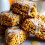 A plate stacked with golden-brown pumpkin scones recipe drizzled with white glaze, with a cup of tea and a teapot in the background.