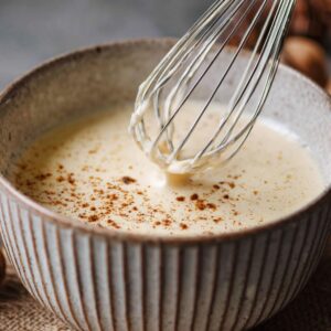 Béchamel sauce in a ceramic bowl with a whisk, dusted with ground nutmeg, surrounded by whole nutmeg and milk jar.