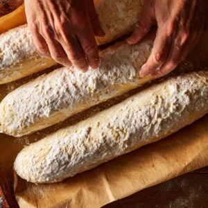 Close-up of hands shaping long dough logs dusted with flour on a parchment-lined baking tray before baking.