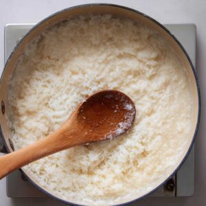 Steamed glutinous rice being combined with coconut mixture in a pot and stirred with a wooden spoon.