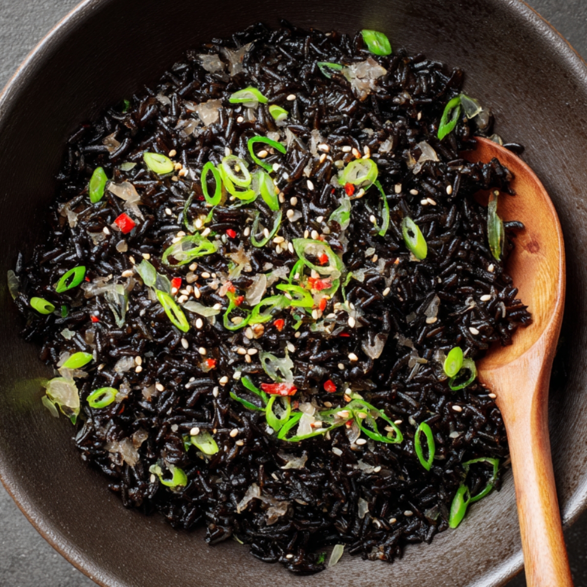 Cooked black rice served in a dark bowl, garnished with sliced green onions, sesame seeds, and red chili flakes, with a wooden spoon on the side.