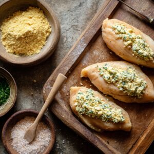 Chicken breasts stuffed with a butter and herb mixture on a wooden board with bowls of breadcrumbs, parsley, and salt.