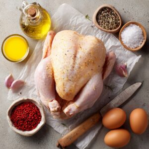 Whole raw chicken with garlic, olive oil, salt, pepper, and eggs laid out on a surface, ready for preparation.