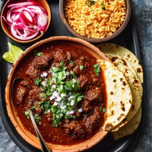 Chili Colorado Bowl: A rich beef stew in red chile sauce, topped with fresh cilantro and diced onion, served with tortillas, Mexican rice, and pickled onions.