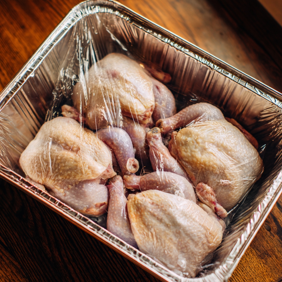 Raw Cornish hens in a foil baking tray, wrapped in clear plastic wrap, ready for preparation.