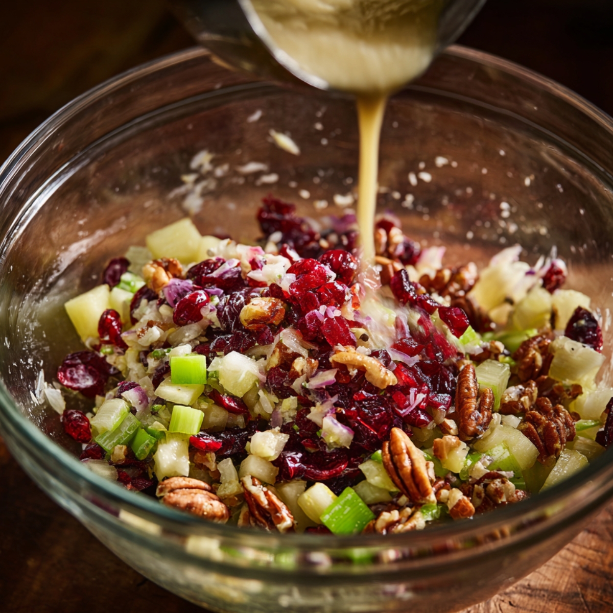 A glass bowl with chopped fruits (likely apples and celery) and cranberries, with nuts being added, followed by a pour of liquid dressing.