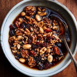 A bowl of mixed nuts, including walnuts, almonds, and peanuts, combined with dried cranberries and a dark syrup.