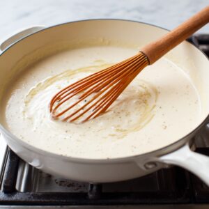 Best Creamy White Chicken Enchiladas Recipe 21 Image of a whisk stirring white sauce in a pan — no visible text present.