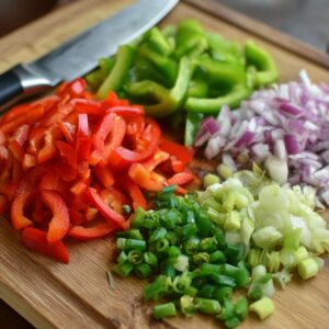 A cutting board with sliced red and green bell peppers, chopped onions, minced green chilies, and chopped spring onions with a knife.