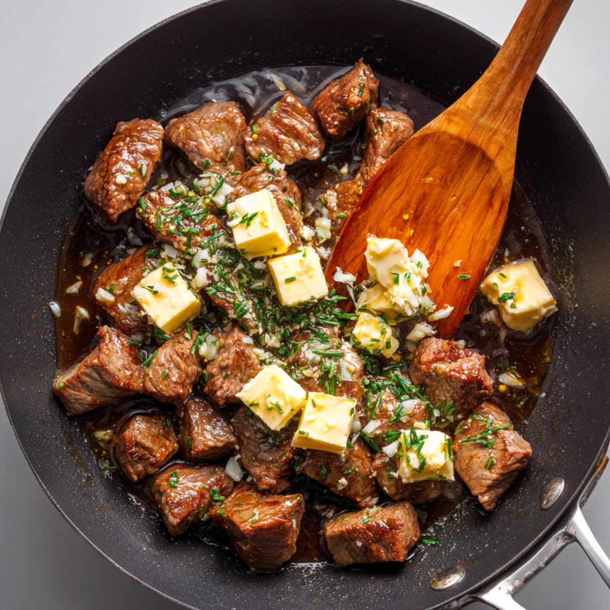 Garlic butter steak bites in pan – Cubes of seared steak in a pan with melted butter, minced garlic, and fresh herbs, being stirred with a wooden spatula.