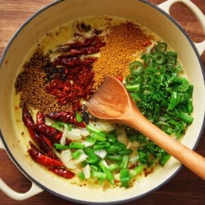 A large skillet with dry spices (mustard seeds, dried red chilies, chili powder) along with onions and fresh green herbs.