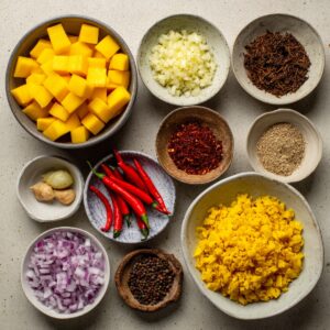 Small bowls containing prepped ingredients: diced mango, chopped onion, garlic, red chilies, ginger, and ground spices.