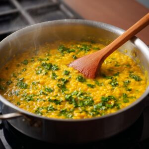 Cook the Dal – A pot of simmering yellow lentils (dal) with chopped green herbs on top, being stirred with a wooden spoon.