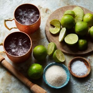 Prep setup with two copper mugs filled with ice, a cutting board with halved and sliced limes, coarse sugar, salt, and a muddler.