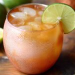 Copper mug Moscow Mule Recipe filled with ice and topped with a lime wedge, surrounded by fresh limes and peaches on a wooden board.