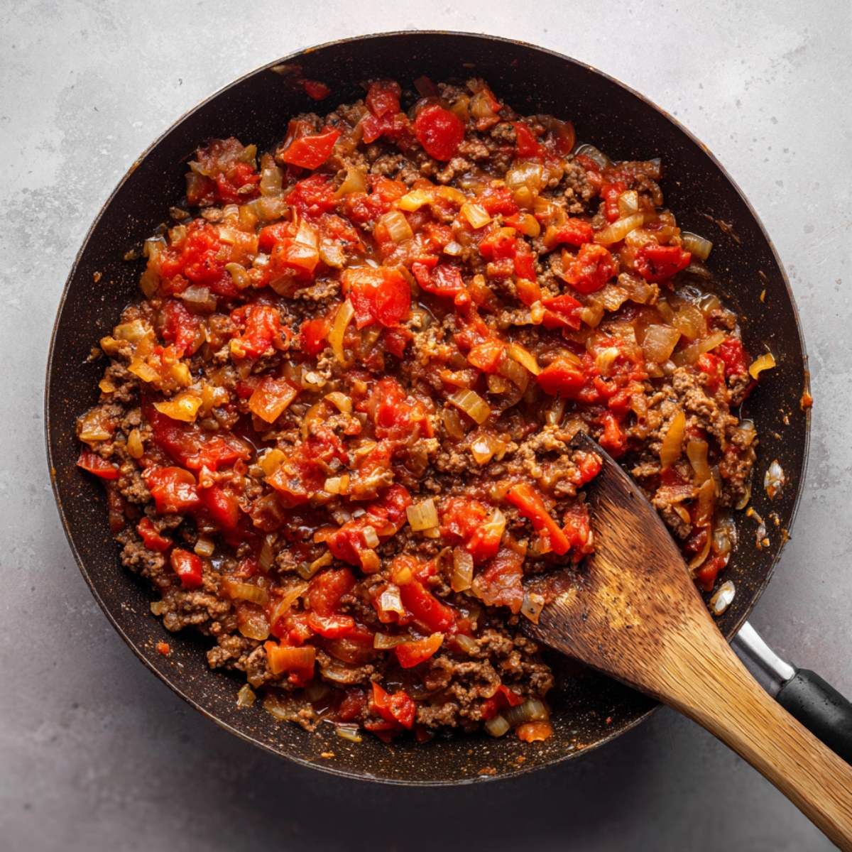 Healthy Moussaka Recipe 13 A skillet filled with ground lamb sauce cooked with onions and tomatoes, with a wooden spatula resting inside.