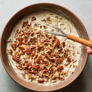 Mixing bowl with nougat mixture and toasted pecans, being folded in by hand with a whisk.