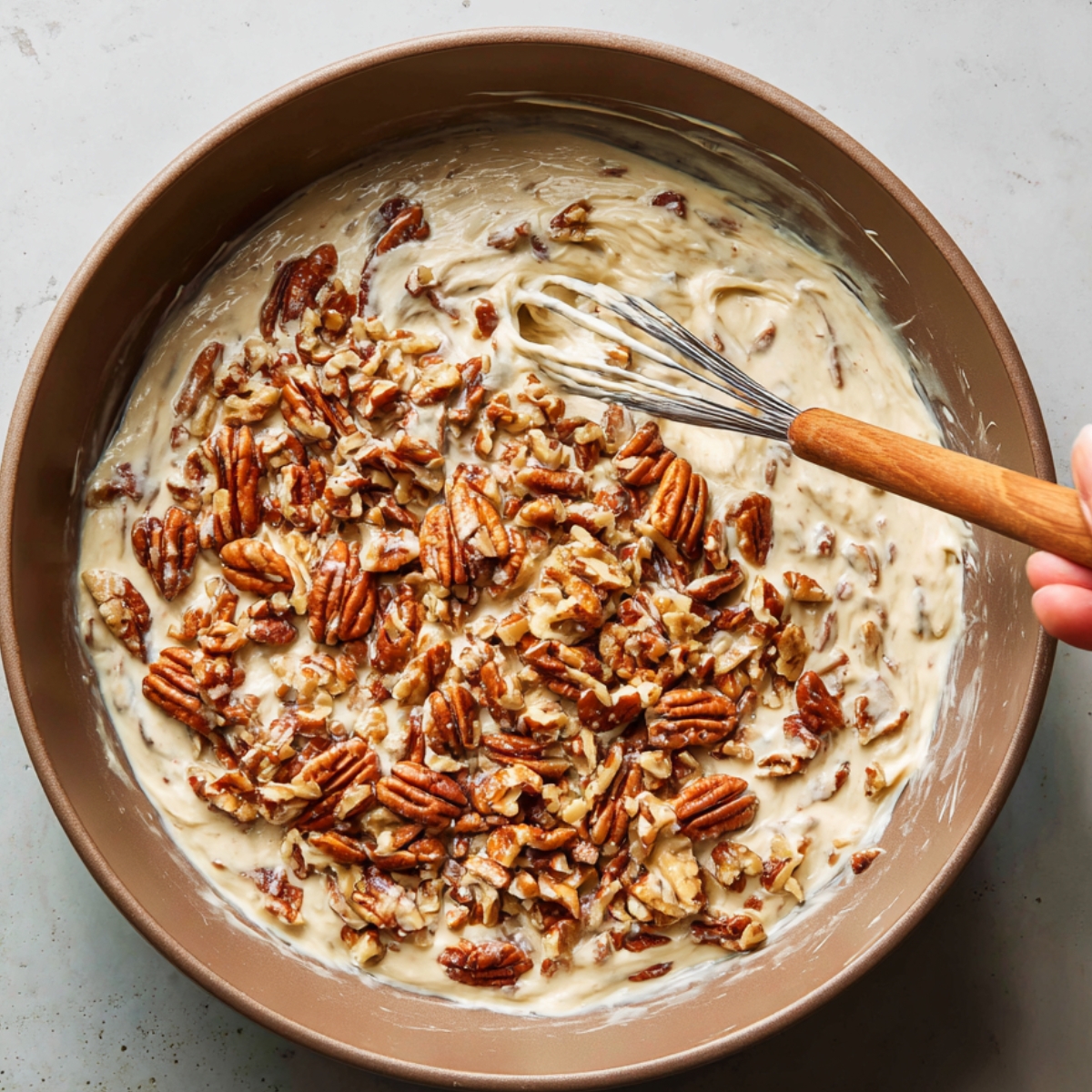Mixing bowl with nougat mixture and toasted pecans, being folded in by hand with a whisk.