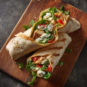 Two grilled wraps placed on a wooden board. Cut open to reveal chicken, lettuce, tomatoes, and creamy dressing. Seasoning sprinkles add a rustic, appetizing touch.