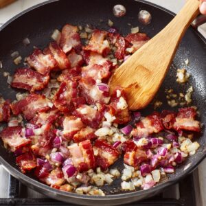 Bacon and chopped onions sizzling in a frying pan being stirred with a wooden spatula.
