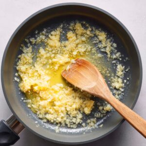 Minced garlic being sautéed in melted butter in a frying pan, stirred with a wooden spoon.