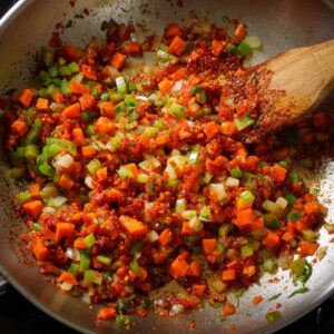 A sauté pan with chopped onions, carrots, and celery cooked with tomato paste, stirred with a wooden spoon.