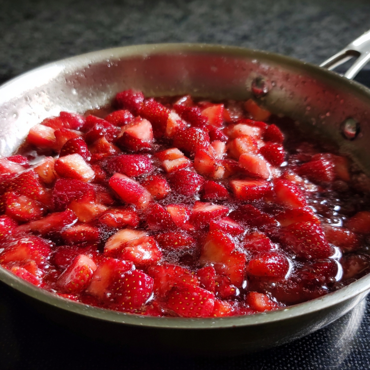 Strawberry sauce cooking: Diced strawberries simmering with sugar in a stainless steel pan, turning into a thick sauce.