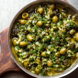 A metal bowl with green olives, capers, parsley, garlic, and olive oil mixed together.