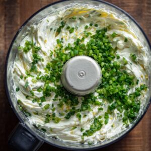 A food processor containing a creamy white mixture (cream cheese base) topped with finely chopped green onions.
