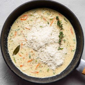 Easy Bacon Cheddar Gnocchi Soup Recipe 20 Roux with flour in creamy soup base – A skillet of simmering broth and vegetables with a large mound of flour and thyme sprigs ready to be whisked in.