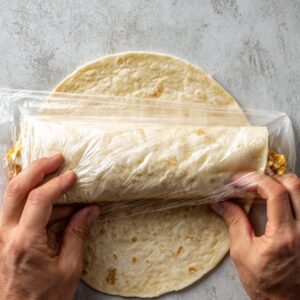 Hands folding a tortilla wrap tightly in plastic wrap to secure the filling inside.
