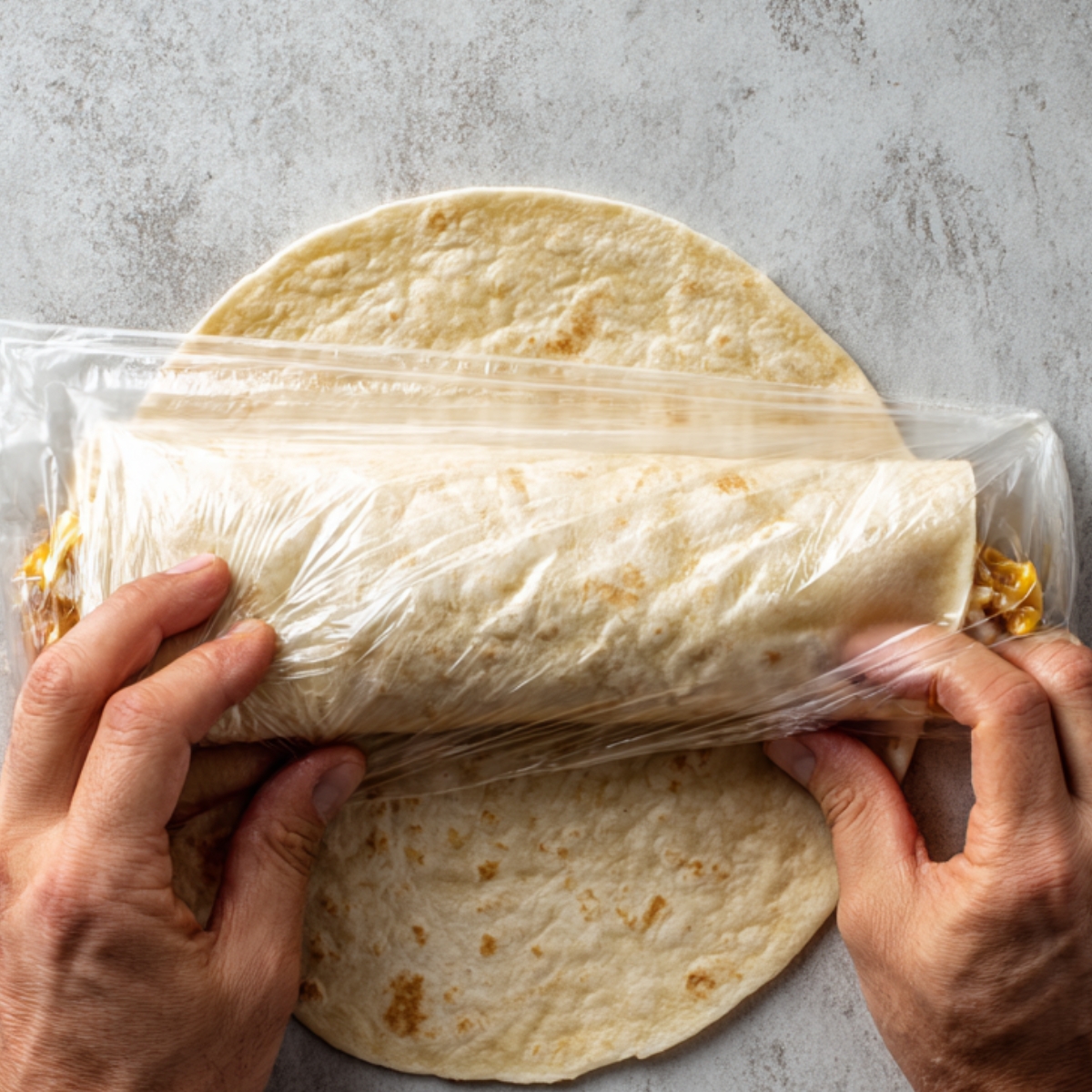 Hands folding a tortilla wrap tightly in plastic wrap to secure the filling inside.