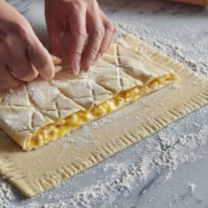 Hands folding pastry dough over a creamy filling on a floured surface with a rolling pin in the background.