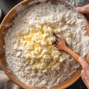 A wooden bowl with flour and small cubes of butter being mixed together with a wooden fork.