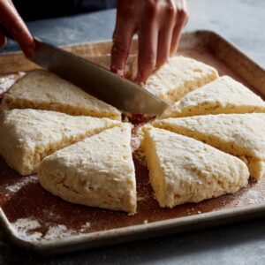 A person using a large knife to cut a round of dough into triangular wedges on a floured baking sheet, preparing scones.