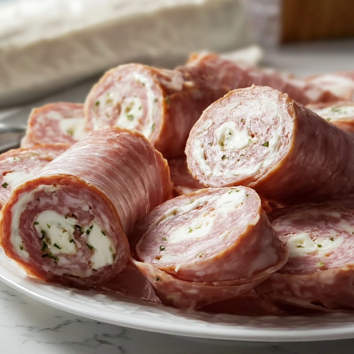 A white plate filled with rolled salami slices stuffed with a creamy cheese and herb mixture, neatly arranged on a marble countertop.