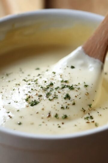 Creamy béchamel sauce recipe in a white bowl with a wooden spoon, topped with fresh herbs and black pepper.