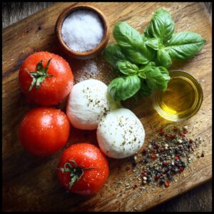 Raw ingredients for a Caprese salad on a wooden board: whole tomatoes, mozzarella balls, fresh basil, olive oil, sea salt in a small bowl, and a mix of pepper and herbs.