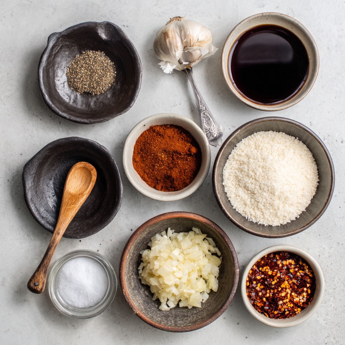 An assortment of small bowls containing spices and ingredients: black pepper, paprika powder, salt, diced onion, Worcestershire sauce, grated Parmesan cheese, red pepper flakes, and a garlic bulb.