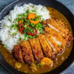 A bowl of Japanese Katsu Curry Recipe rice topped with sliced crispy chicken cutlet, curry sauce with carrots, and garnished with sliced green onions and red chili peppers.