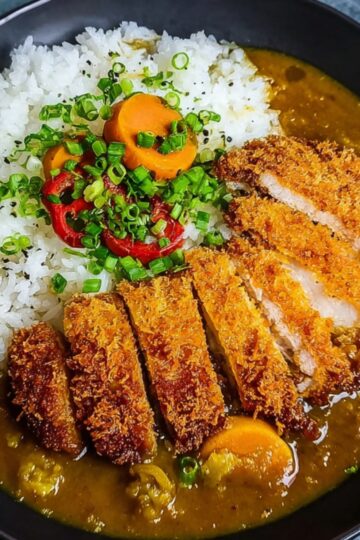 A bowl of Japanese Katsu Curry Recipe rice topped with sliced crispy chicken cutlet, curry sauce with carrots, and garnished with sliced green onions and red chili peppers.