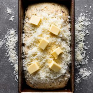 Unbaked meatloaf topped with cheese and butter – Raw meatloaf in a loaf pan, covered with shredded Parmesan and dotted with cubes of butter.