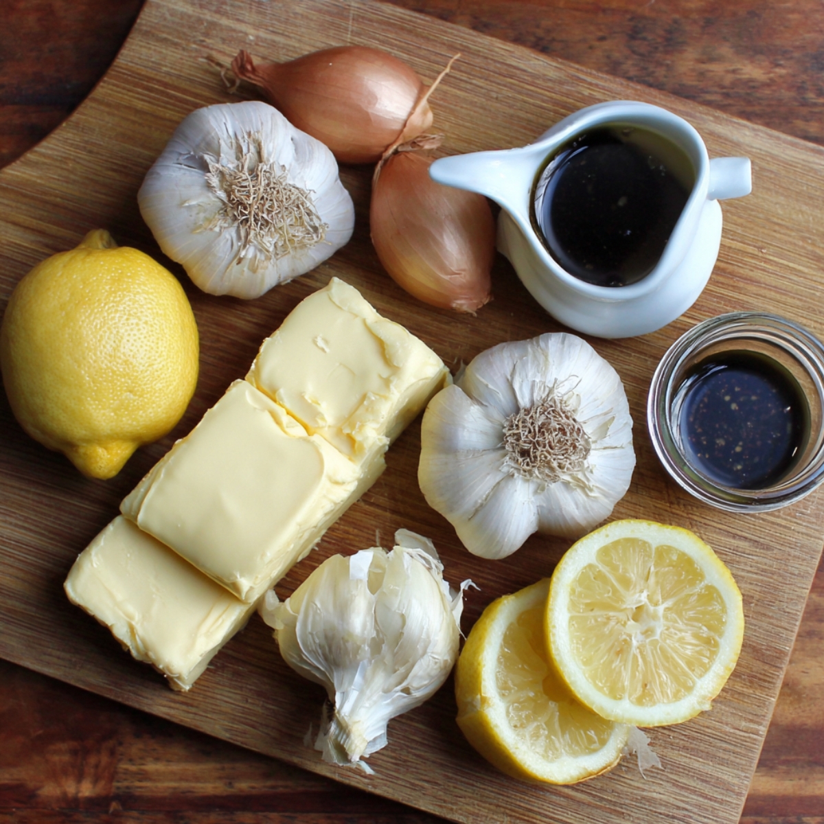 Fresh garlic, shallots, butter blocks, lemon, and small jars of Worcestershire sauce on a wooden board.