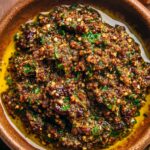 A small white ramekin filled with Tapenade Recipe (a blend of finely chopped olives, capers, garlic, and herbs) served with toasted garlic bread slices.