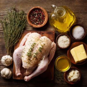 Raw whole turkey on a wooden board surrounded by garlic, thyme, butter, olive oil, salt, and pepper