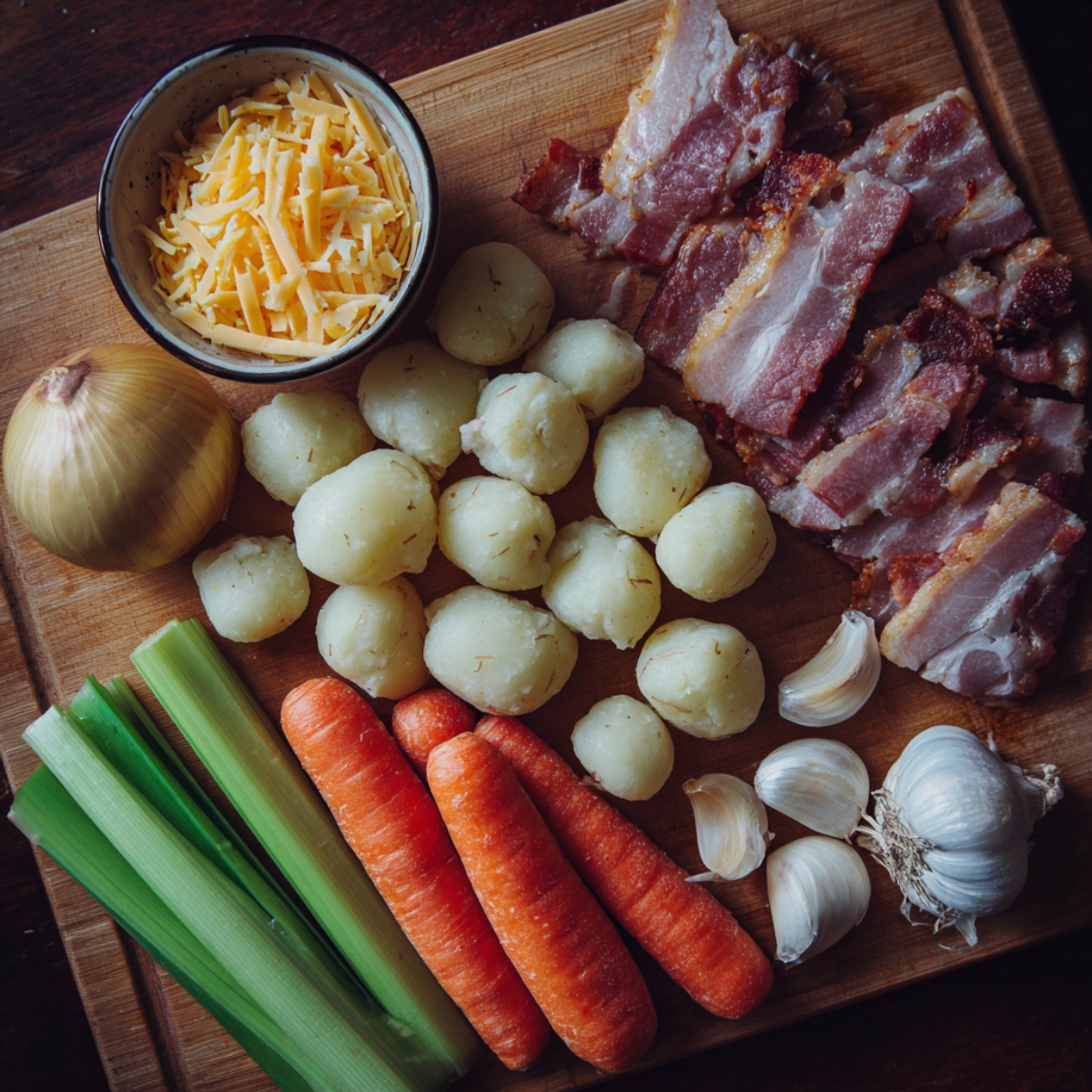 Easy Bacon Cheddar Gnocchi Soup Recipe 10 Soup base ingredients on cutting board – Strips of cooked bacon, potato gnocchi, shredded cheddar, carrots, celery, garlic, and onion laid out together.