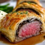 A close-up of a Beef Wellington Recipe, showing a slice with perfectly cooked beef inside, wrapped in golden-brown puff pastry. It’s served with mashed potatoes, green beans, and a drizzle of sauce.