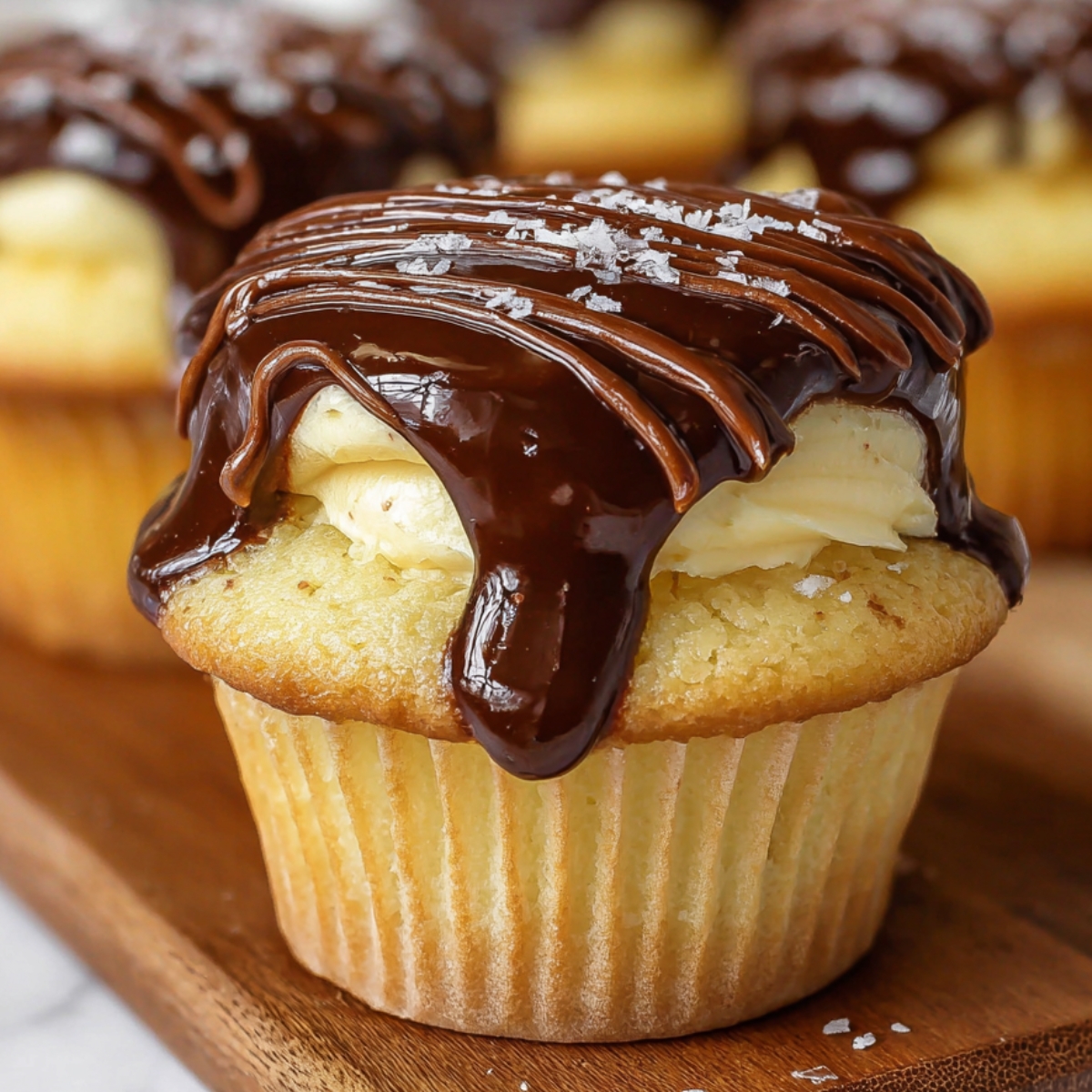A close-up of a vanilla cupcake topped with thick, glossy chocolate ganache, sprinkled with flakes of sea salt, sitting on a wooden board.