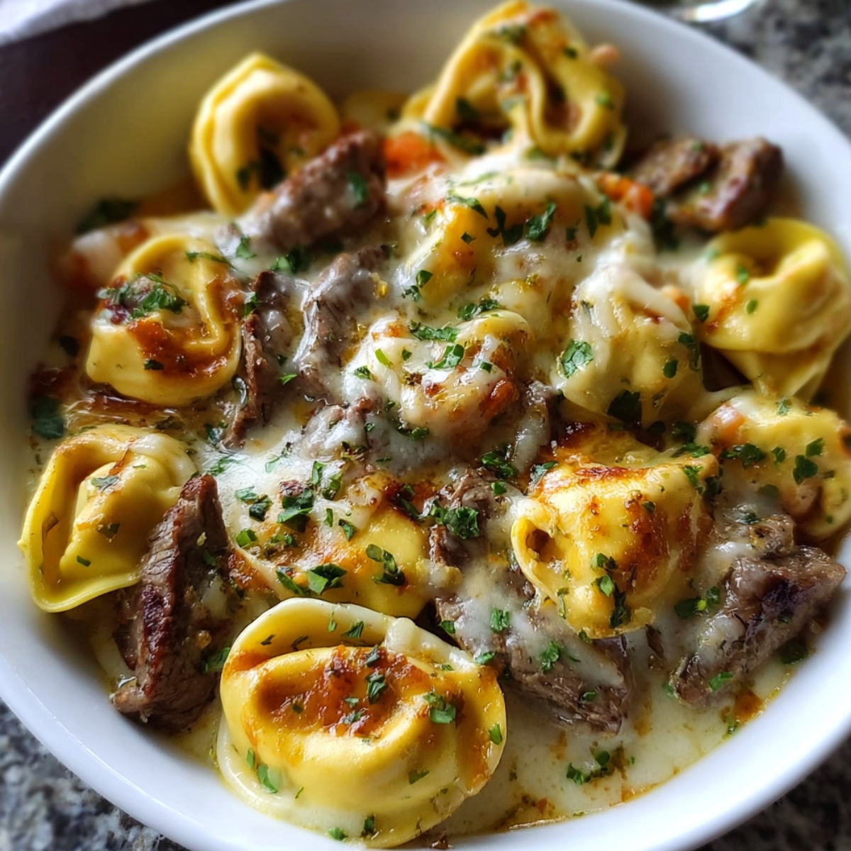 Best Cheesesteak Tortellini Provolone Sauce Recipe 12 A bowl of tortellini pasta mixed with tender steak pieces, smothered in creamy provolone sauce, and sprinkled with parsley.