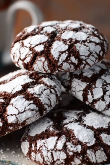 A plate with several chocolate crinkle cookies dusted with powdered sugar, with a cup of coffee in the background.